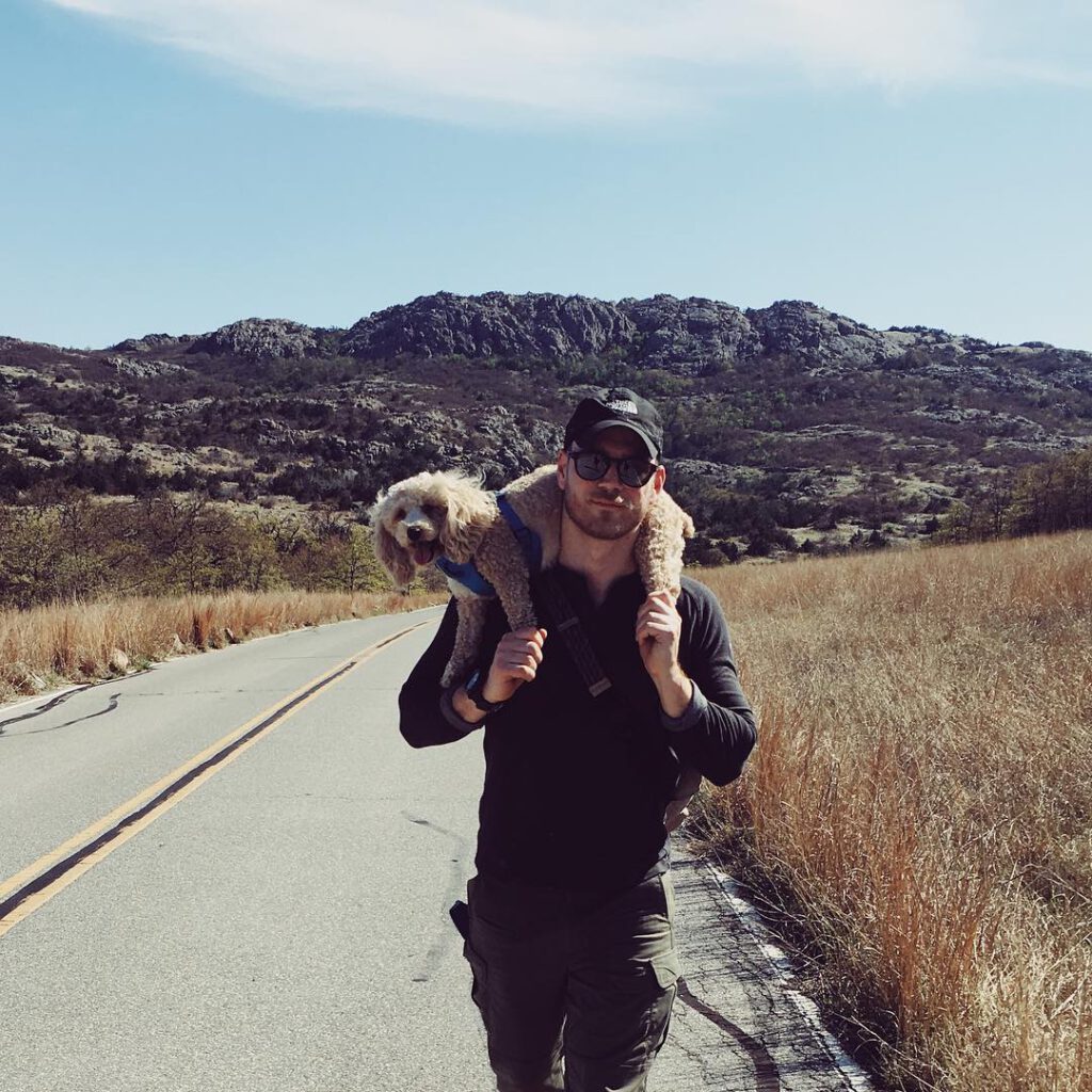 Richard is pictured facing the camera walking down a road with a small dog on his shoulders in front of low mountains.